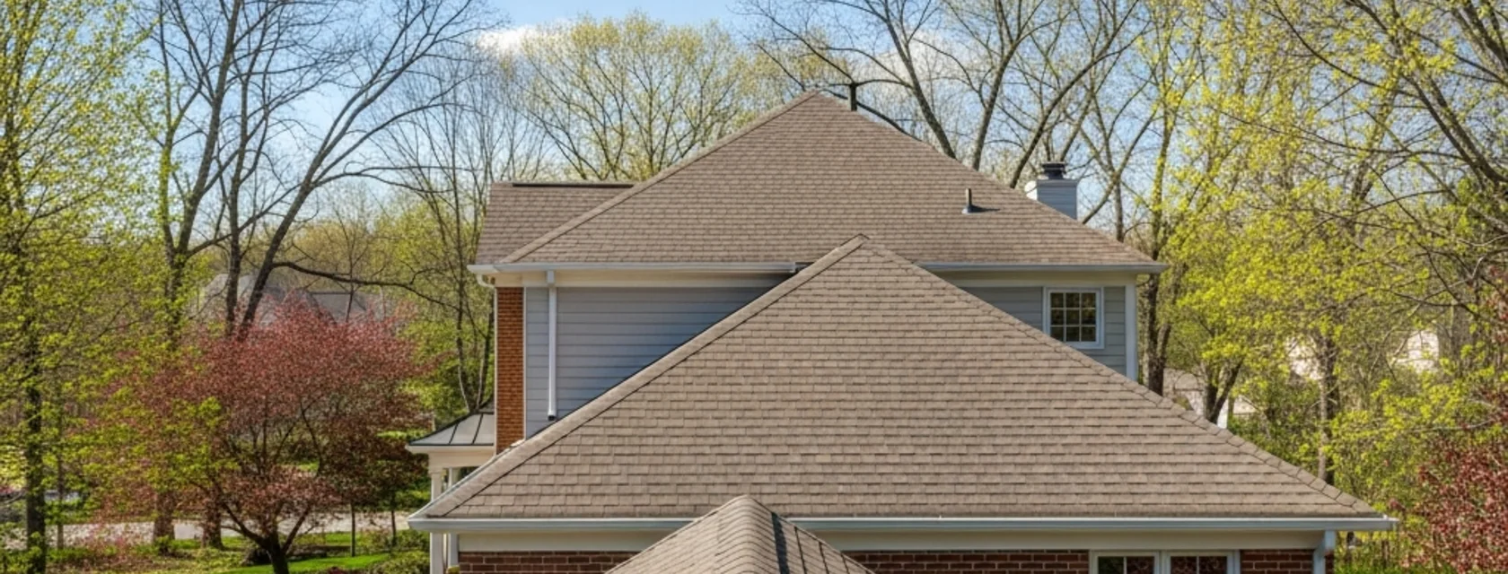 Shingle roof with two hipped slopes, a chimney, white gutters and a downspout, surrounded by trees with new spring leaves