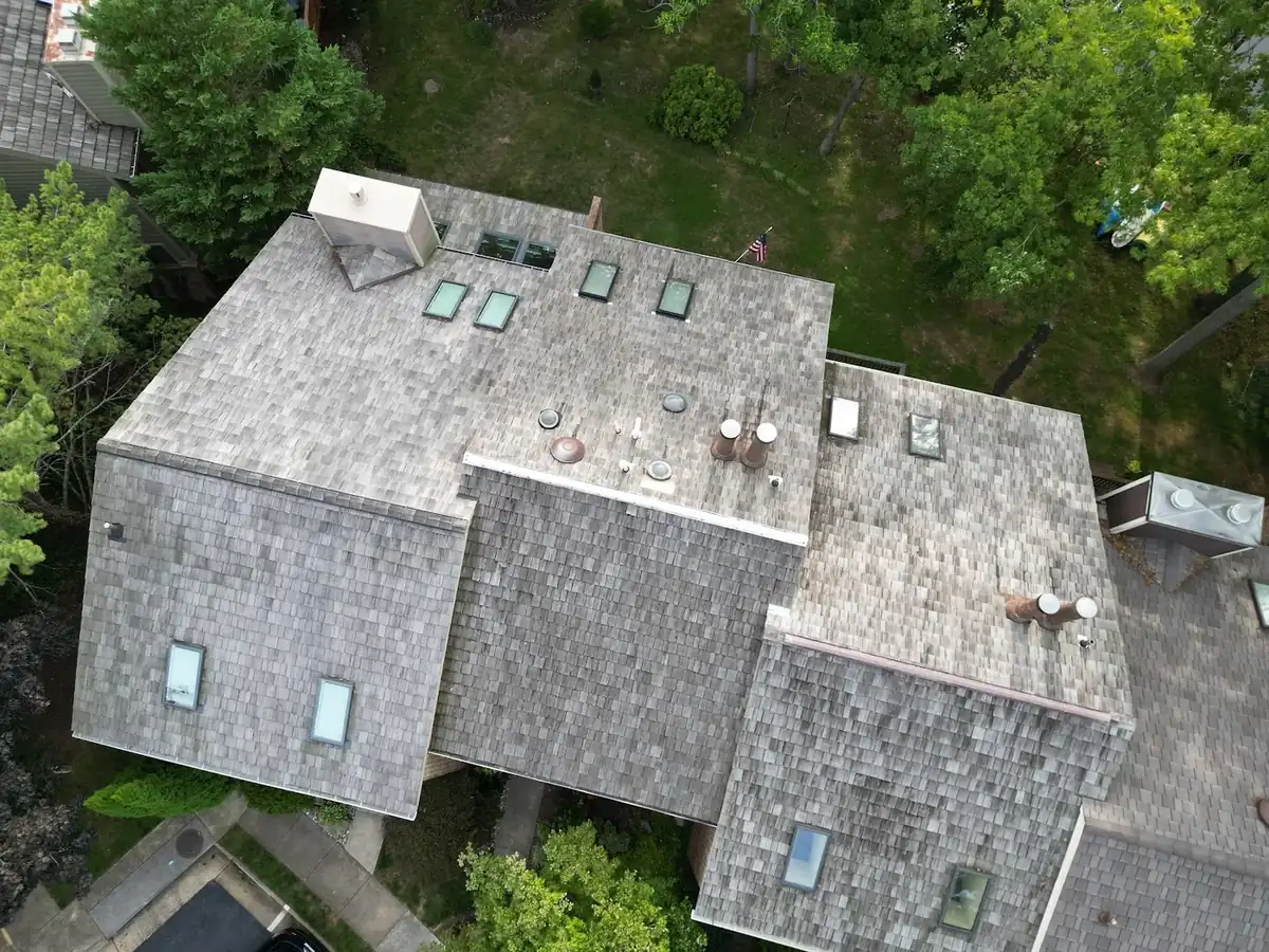Aerial view of a gray cedar shake roof with several skylights, vent pipes and two chimney caps, surrounded by trees