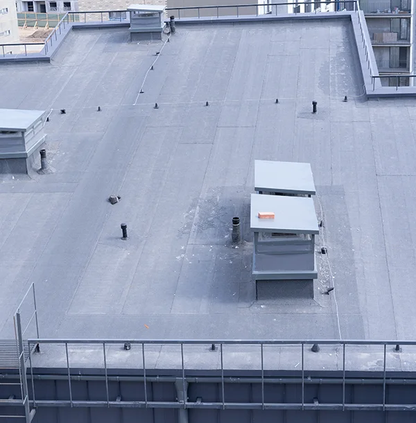 Flat membrane roof seen from above with vent pipes, two rooftop units and a metal railing along the parapet