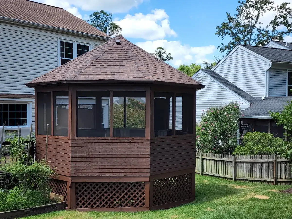 Octagonal screened gazebo with a brown shingle roof in a backyard next to two houses
