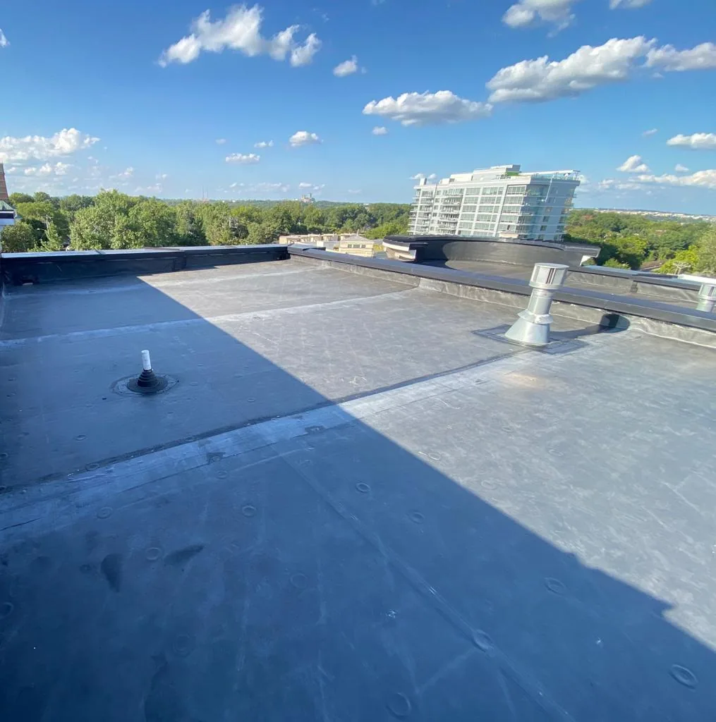Dark single-ply flat roof with a pipe vent, a metal vent cap and a raised parapet edge, with a tall apartment building in the distance