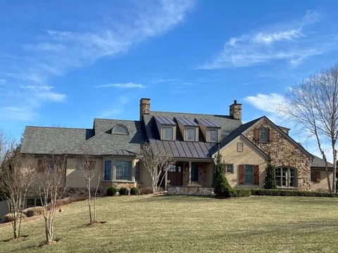 Large stone and stucco house with a dark shingle roof and standing-seam metal panels on the entry roof and dormer tops