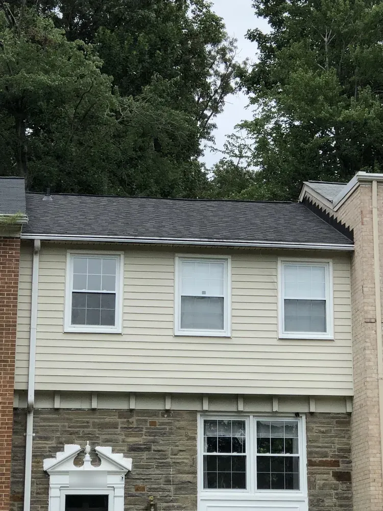 Two-story attached house with a dark asphalt shingle roof, beige siding, brick and stone facing and tall trees behind it