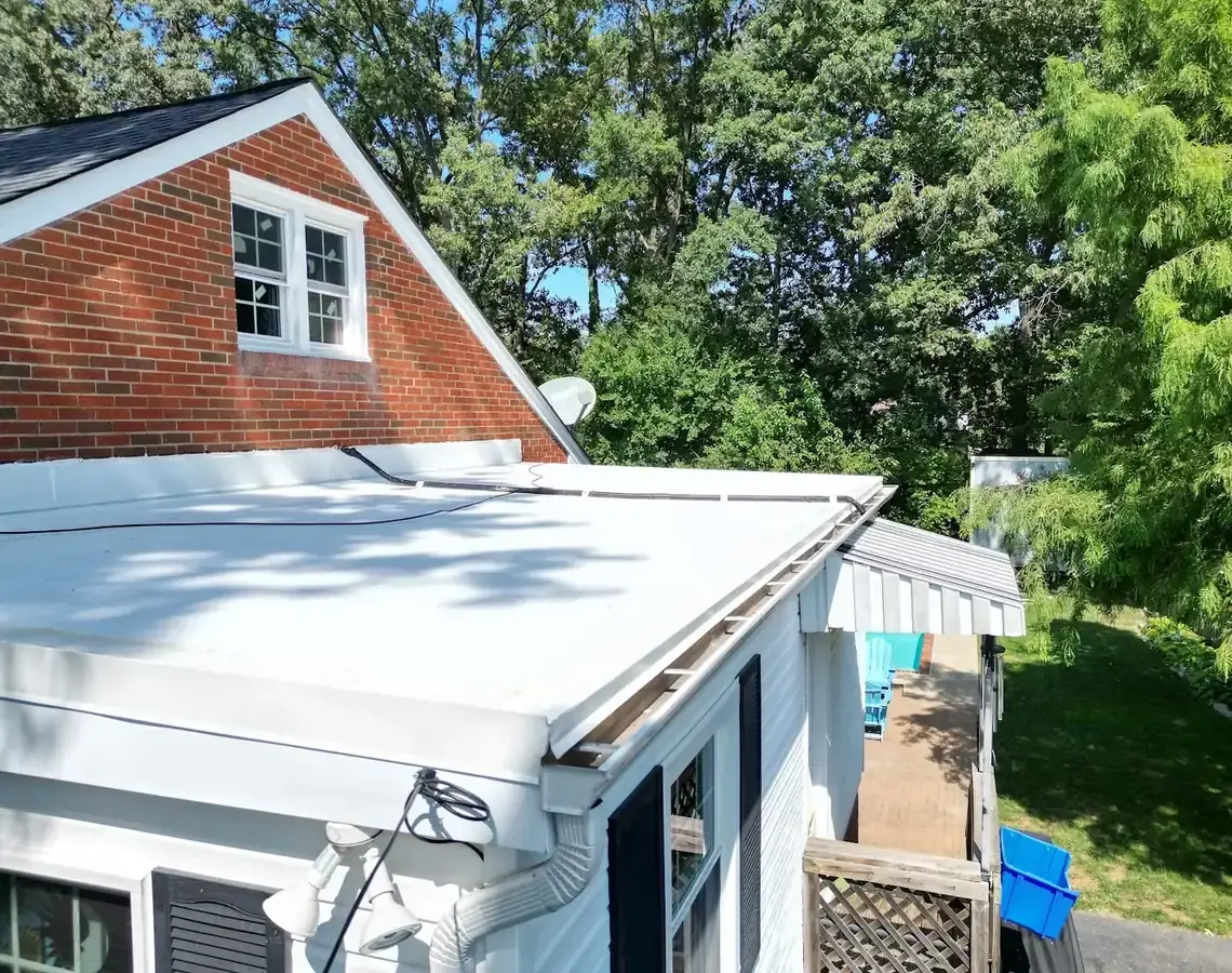 Light-colored flat roof over a house addition beside a brick gable with a white-framed window, with trees behind
